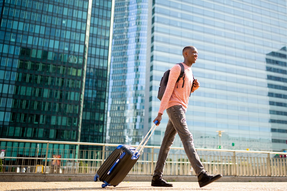 Full body side of young black man walking with suitcase in city Glücklicher Geschäftsreisender, der mit Koffer durch die Stadt läuft