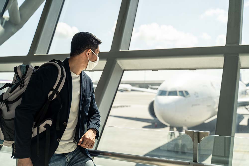 Man with mask on carrying a backpack looking at plane from inside an airport