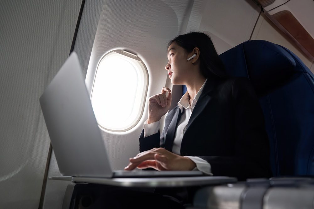 Belle femme d'affaires asiatique assise près de la fenêtre dans un avion écoutant de la musique avec des écouteurs tout en travaillant avec un ordinateur portable femme travaillant depuis un siège de cloison